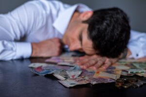 A man in a white shirt rests his head on a table covered with Argentine pesos and coins.